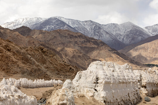 View of a row of white stupas set against the rugged, snow-dusted Himalayan mountains in the high-altitude desert landscape of Leh, Ladakh, India.