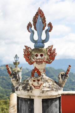 View of the intricate skull-themed decorative finial atop a pillar at Namobuddha Monastery, set against a backdrop of lush mountains in Simalchaur Syampati, Bagmati Province, Nepal.