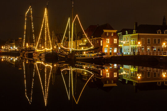 View of historic Dutch buildings and sailboats decorated with festive fairy lights reflecting in the calm canal water at night in Weesp, North Holland, Netherlands.