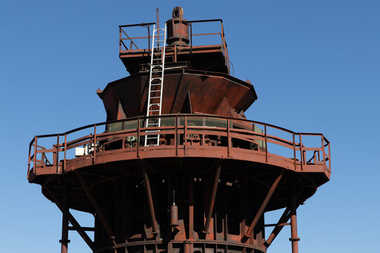 View of the rusty industrial blast furnace structure with metal railings and a ladder against a clear blue sky in Velsen-Noord, North Holland, Netherlands.