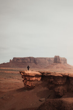 Arizona, USA - 22 March 2025: View of a person standing on a rocky ledge overlooking the iconic sandstone buttes and mesas of Monument Valley under an overcast sky.
