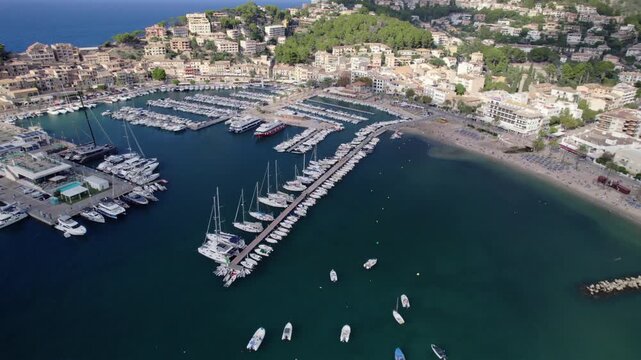 Drone shot captures Port de Soller&rsquo;s marina, luxury yachts, turquoise waters, sandy beach, and hillside buildings on Mallorca island under bright natural daylight.