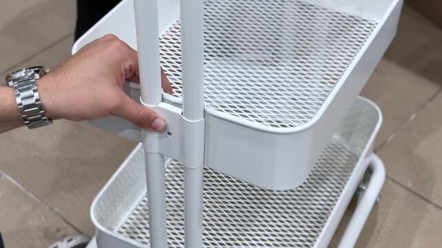 Close-up of hands assembling a white metal utility cart indoors. The person connects the steel frame components over a tiled floor.