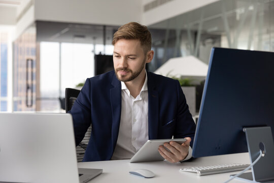 Busy young man fintech professional or digital consultant multitasking across laptop tablet and pc managing cross device business operations comparing information in multi screen corporate environment
