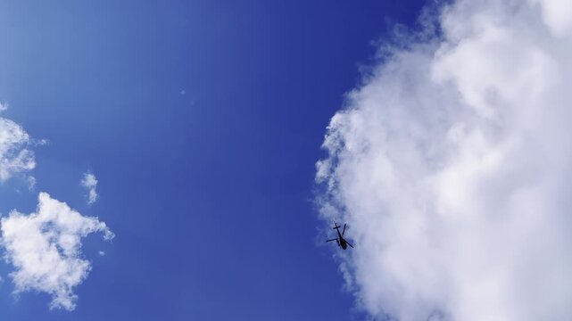 Helicopter silhouette hovering near large white cumulus cloud against deep blue sky, shot from ground level looking upward, bright natural daylight, aerial perspective with minimal cloud coverage
