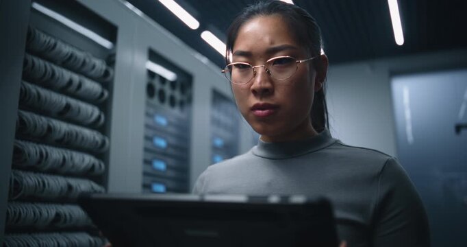 Young Female Data Center Specialist Studies Tablet With Concern While Monitoring Server Infrastructure. Focused Expression Suggests Technical Analysis, Diagnostics and Network Issue in Facility.