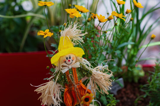 Happy small handmade scarecrow doll with yellow hat and straw hair standing among bright yellow flowers. Close up of the decorative figurine in a garden setting with green stems and soft background.