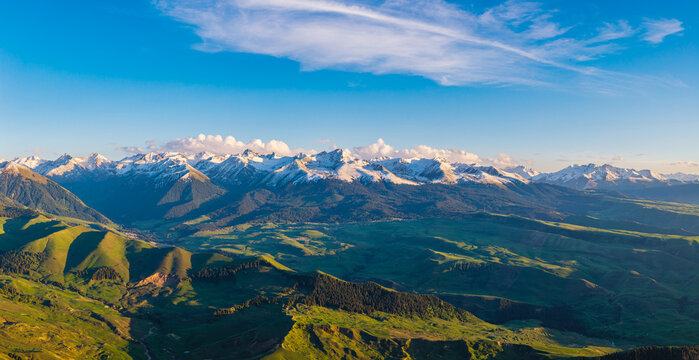 Aerial panorama of snow-capped mountain ridges and undulating green valleys under a clear blue sky in Kalajun Grassland, Ili, Xinjiang, China
