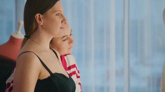 Two women stand side by side, looking out a window. The woman in black appears calm and thoughtful. The woman in red stripes shows concern