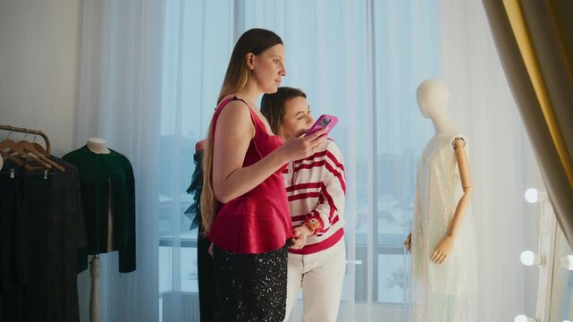 Mother and daughter browse clothes in boutique. Girl looks at phone, mother points to outfit. They smile and walk through store. Footage fits retail, fashion, family, shopping themes