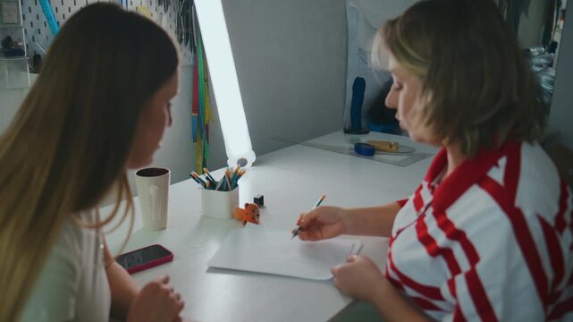 Two women collaborate at a desk, one draws while the other watches. Artist focuses intently, sketching with pencil. Young woman observes closely, holding phone
