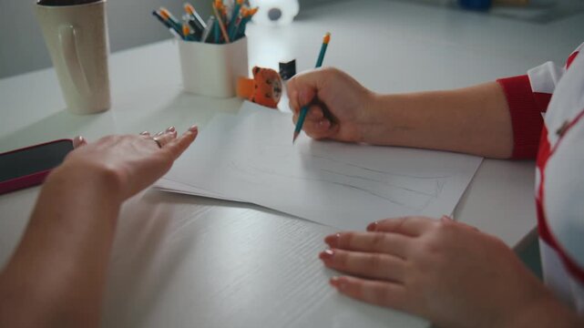 Two people sketching on paper at white desk. One holds pencil, other guides design. Hands show focus and collaboration. Office tools nearby indicate creative work. Ideal for design, architecture
