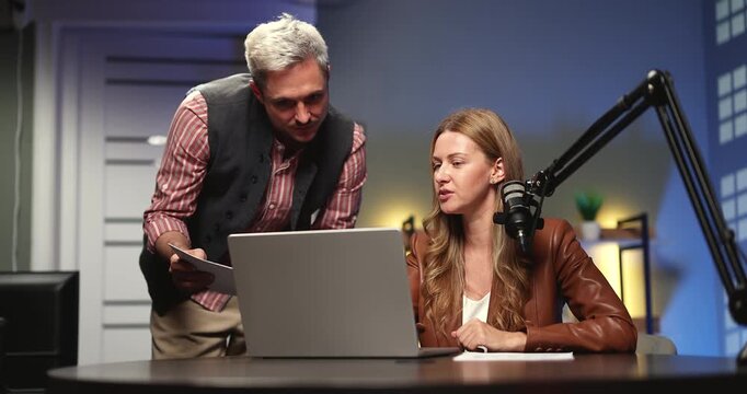 Media professional or presenter consulting with producer or technical assistant planning recording session editing narrative content using laptop paper scripts during pre production in modern studio