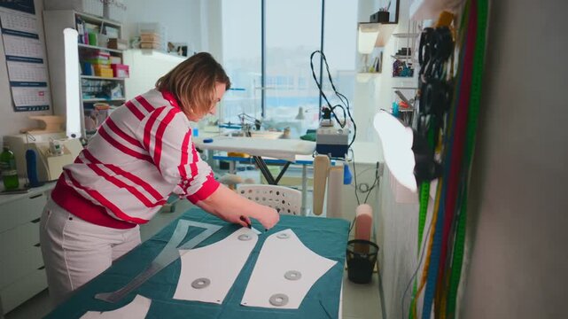 Woman carefully arranges pattern pieces on cutting table. Bright, organized sewing space with natural light. Focused expression shows dedication to craft. Ideal for fashion, tailoring, or DIY projects