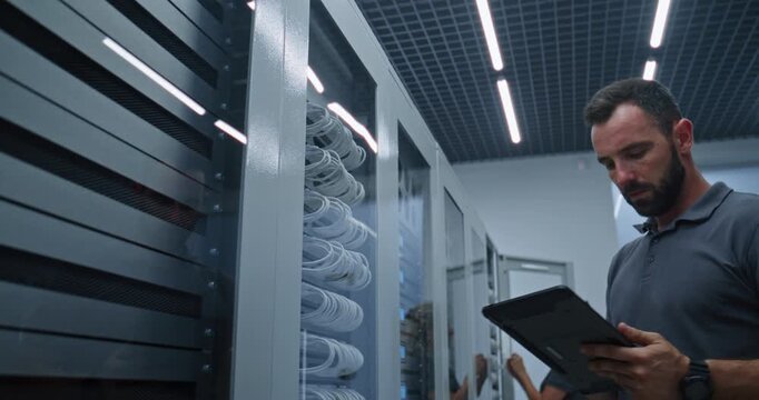 IT Technician Monitors Server Performance on Tablet While Checking Network Ports, Cable Connections, Documenting Infrastructure Status and Troubleshooting Connectivity Inside Enterprise Server Room.