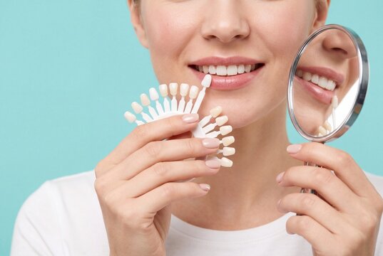 Close-up of smiling woman matching tooth shade with dental color guide and hand mirror, teeth whitening and cosmetic dentistry concept on turquoise studio background