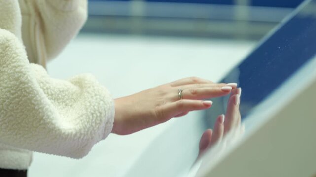 Person touches a modern interactive display screen. Their hand shows rings and soft white sleeves. The background is blurred with blue and white tones. Human interaction with technology indoors