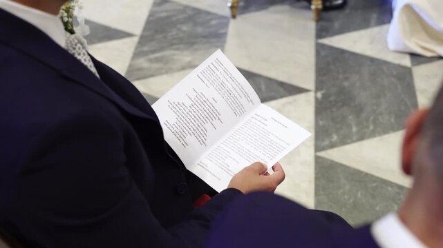 A seated man in a navy suit holds and reads an open printed booklet above his lap, with a geometric stone floor and another suited figure nearby.