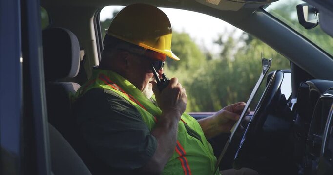 Utility worker talks on handheld radio while reviewing clipboard inside work vehicle