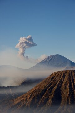 Vertical view of Mount Semeru erupting with billowing ash cloud above Mount Batok, blue sky and morning haze, East Java, Indonesia