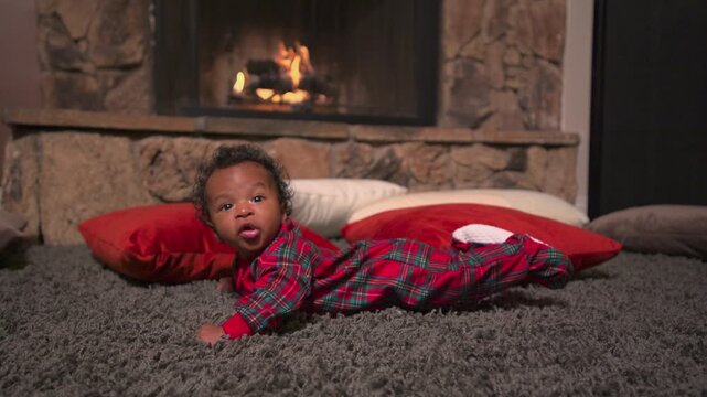 An excited infant in a red tartan sleeper smiles open mouthed during tummy time on a gray shag rug. Red and white floor pillows lie before a stone fireplace with burning logs. Cozy Christmas evening.