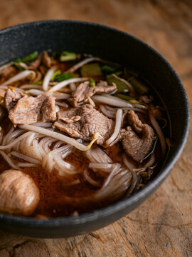 Close-up of Thai boat noodle soup with sliced beef, meatball, rice noodles, bean sprouts and green onion in rich dark broth, served in a black bowl on rustic wooden table
