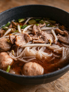 Close-up of Thai boat noodle soup with sliced beef, meatball, rice noodles, bean sprouts and green onion in rich dark broth, served in a black bowl on rustic wooden table