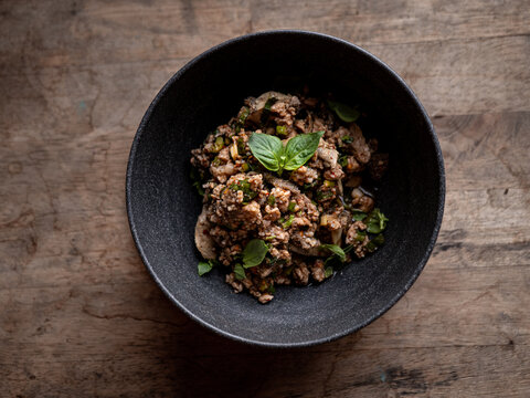 Thai spicy minced pork larb salad topped with fresh basil leaves, chili and herbs, served in a black bowl on a rustic wooden table, top view of northern Thai food