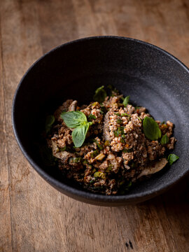 Thai spicy minced pork larb salad topped with fresh basil leaves, chili and herbs, served in a black bowl on a rustic wooden table, top view of northern Thai food