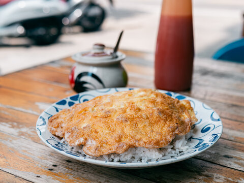 Thai-style fried omelette over steamed white rice on a blue and white ceramic plate, rustic weathered wooden table with a rooster bowl in the background