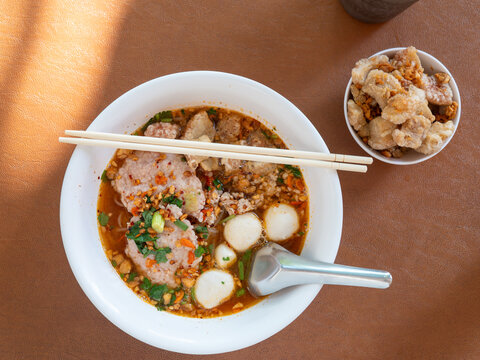 Thai spicy tom yum noodle soup with pork slices, minced pork, fish balls, crispy pork rinds, peanuts and herbs in a white bowl with chopsticks and spoon, street food lunch