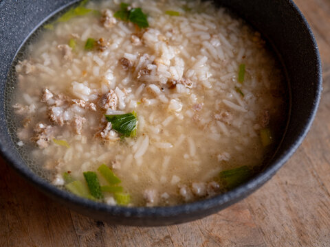 Thai rice soup with minced pork, bok choy and green onion in clear savory broth served in a dark ceramic bowl on a rustic wooden table, comforting breakfast dish