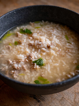 Thai rice soup with minced pork, bok choy and green onion in clear savory broth served in a dark ceramic bowl on a rustic wooden table, comforting breakfast dish