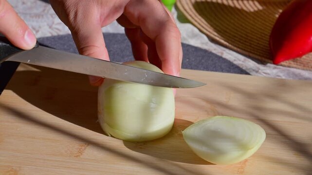 a person using a sharp kitchen knife to slice a fresh, peeled white onion on a wooden cutting board in bright natural sunlight
