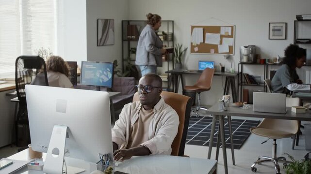 African American young adult man typing on desktop computer and thinking deeply in multiethnic office. Perfect for IT technology and corporate workflow projects