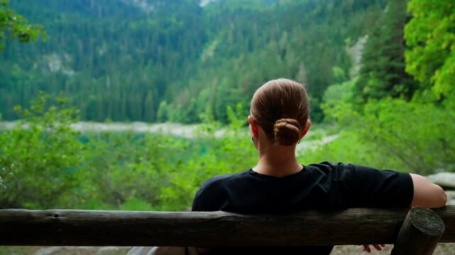 Woman sitting alone on a rustic wooden bench, peacefully admiring a lush green forest valley and distant mountains on a tranquil nature getaway
