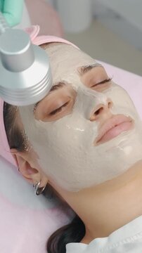 young woman receiving facial treatment with creamy white mask while esthetician applies green led light across