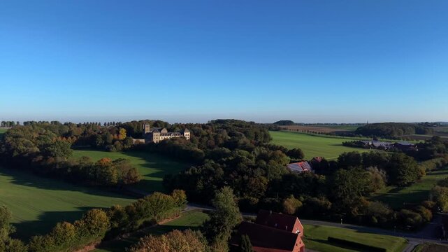 Historic Gerleve monastery sits amid dense autumn woodland, red-roofed farmhouse and rolling green pastures stretching across tranquil rural German hillside landscape. Aerial drone wide shot