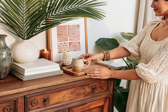 Woman placing ceramic mug on wooden console with plants and decor in bright home interior