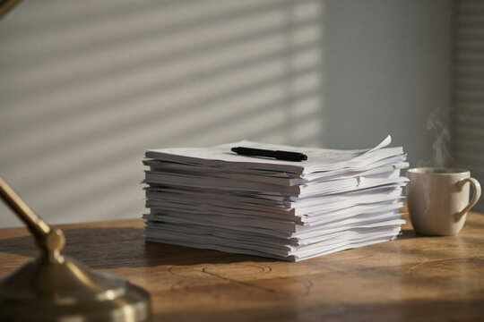 A tall stack of loose paper on a dark wooden desk