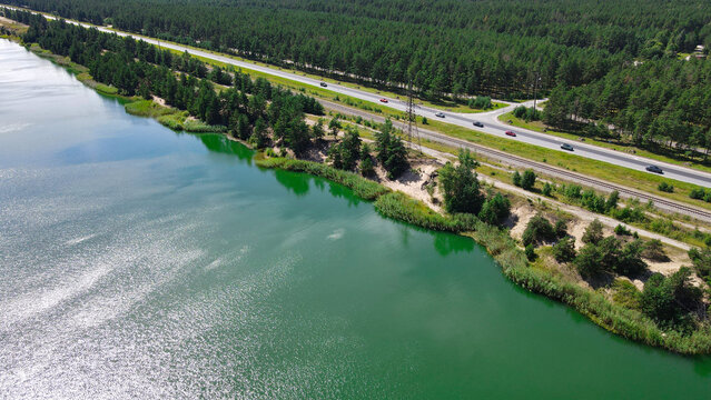 Green lake water reflecting the sunny sky, sitting adjacent to a busy highway with cars and parallel railroad tracks, all framed by a dense pine forest under a clear summer day