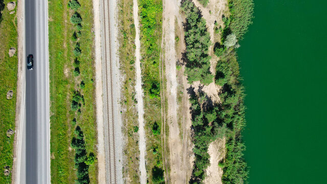 Black car driving on a parallel highway and railroad tracks, separated by green landscape, running alongside a vibrant green lake from a top down aerial perspective