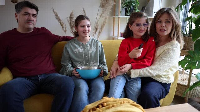 Smiling family sitting together on yellow couch, watching television and holding popcorn bowl while relaxing in bright modern living room during cozy weekend