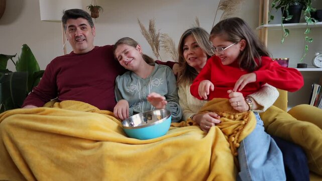 Cheerful family sitting together under a blanket on a yellow sofa, watching an exciting scary movie and getting surprised while spilling popcorn in their living room