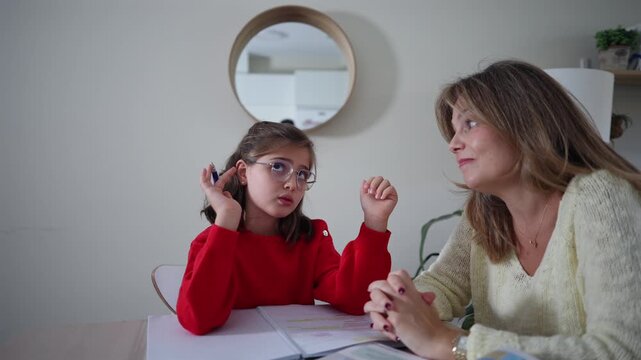 Caring mother listening to her little daughter explaining school exercises, sitting together at a desk during a home education session with textbooks and notebooks