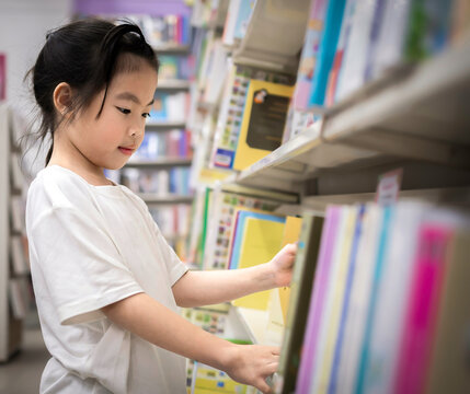 Little girl choosing book in public library room selecting literature for reading.