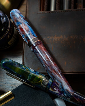Set of luxury colorful acrylic fountain pens next to a vintage pocket watch and leather notebook on a wooden desk.