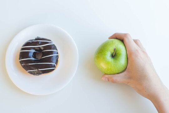 Hand holding a fresh green apple making a healthy choice concept contrasting with a sweet chocolate glazed donut on a plate over a clean white background