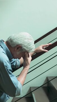 late 50s southeast asian man with white hair suffering headache and dizziness on a home staircase indoors, holding his head in distress for health and aging concepts, vertical video