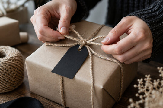 Close-up of hands tying natural twine around a kraft paper gift box with a blank black tag on a rustic wooden table.